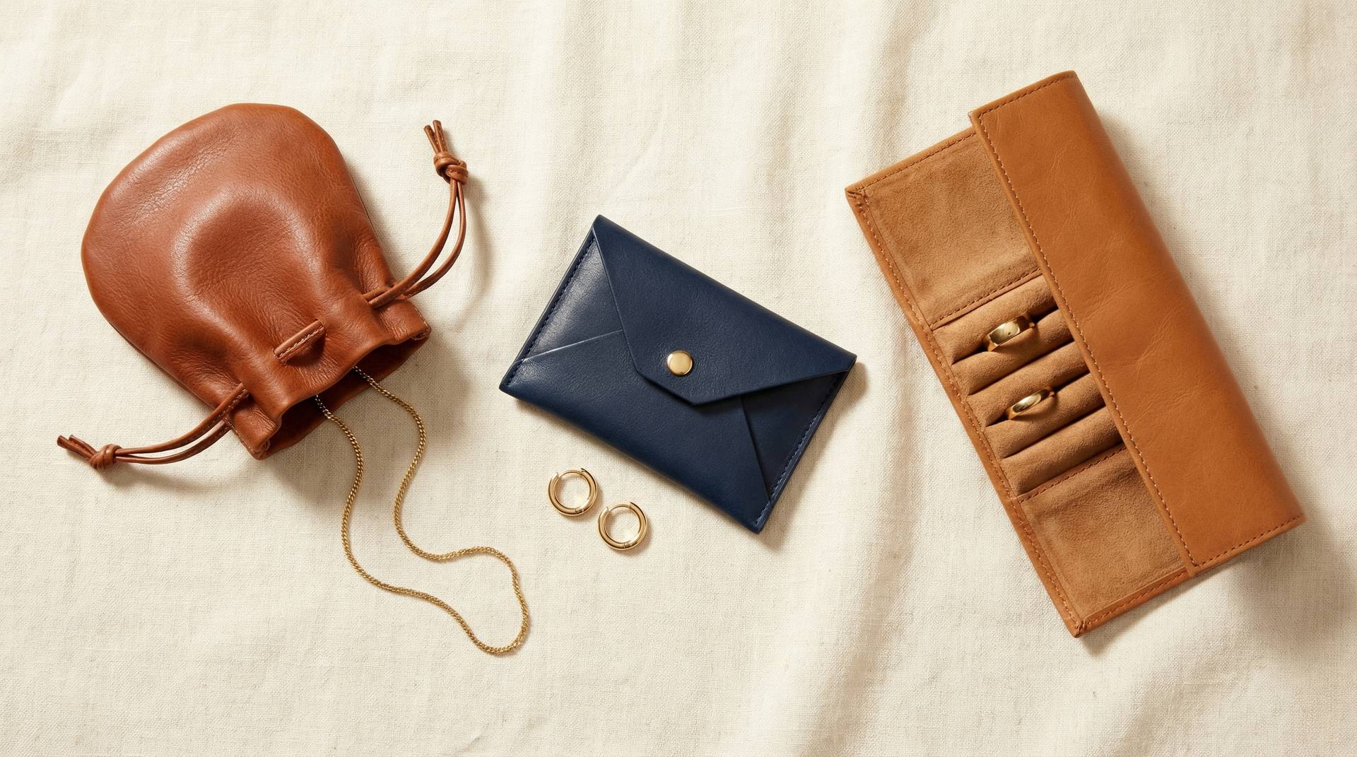 Flat lay of genuine leather jewelry pouches — cognac drawstring, navy snap-closure, and tan envelope on cream linen